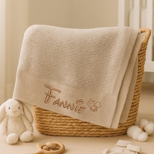 May include: A beige baby blanket embroidered with the name "Fannie" and a bee design, folded in a woven basket. A stuffed bunny, wooden rattle, and baby socks are nearby, suggesting a nursery theme.