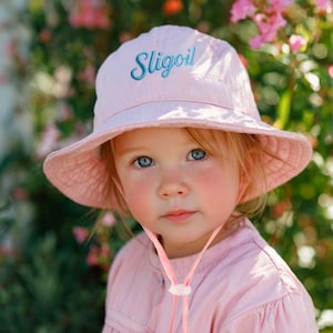 May include: A light pink sun hat with a wide brim and the word "Sligoil" embroidered in blue script. The hat has a chin strap and is worn by a person wearing a pink shirt. The background is a blur of green and pink.