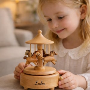 May include: Wooden carousel music box with four carved horses. The carousel has a domed roof and a round base. The name "Luka" is written on the base. The music box is made of light-colored wood.