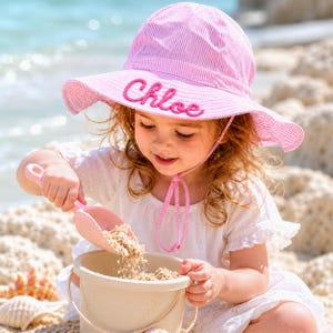 May include: A pink and white striped sun hat with the name "Chloe" embroidered on the front. The hat has a wide brim and a chin strap. The hat is worn by a child playing with a pink shovel and a beige bucket on the beach.