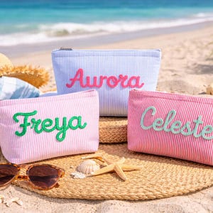 May include: Three personalized cosmetic bags with names embroidered in different colors. The bags are pink and blue with a seersucker stripe pattern. The bags are on a straw mat with sunglasses, seashells, and a beach background.