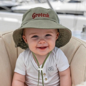 May include: A smiling baby wearing an olive green sun hat with the word "Grotesk" embroidered in red. The baby is also wearing a white onesie with a small design and green drawstring. The baby is sitting on a beige chair.