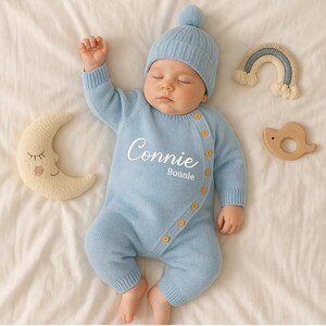 Op de afbeelding: Een slapende baby in een lichtblauwe gebreide romper en bijpassende muts met een pompon. De romper heeft de naam "Connie" erop geborduurd. Gele knopen accentueren de sluiting aan de zijkant. Een maansikkel en een regenboogvormig speelgoed liggen in de buurt.