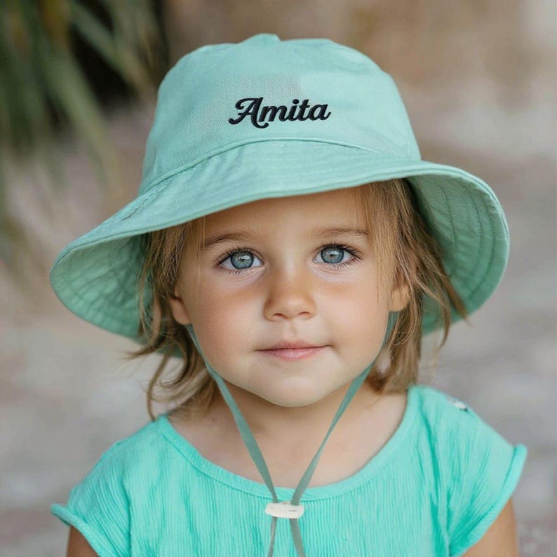 Pu&ograve; includere: Un cappello da sole verde chiaro con il nome "Amita" ricamato in corsivo nero. Il cappello ha un'ampia tesa e un sottogola. La persona nell'immagine indossa un top verde chiaro abbinato.