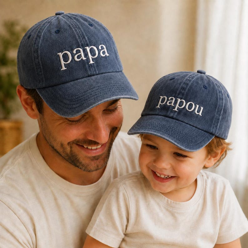 K&ouml;nnte beinhalten: Zwei blaue Baseballkappen aus Denim. Auf der gr&ouml;&szlig;eren Kappe ist das Wort "papa" wei&szlig; gestickt. Auf der kleineren Kappe ist das Wort "papou" wei&szlig; gestickt. Die Kappen werden von einem Mann und einem Kind getragen.