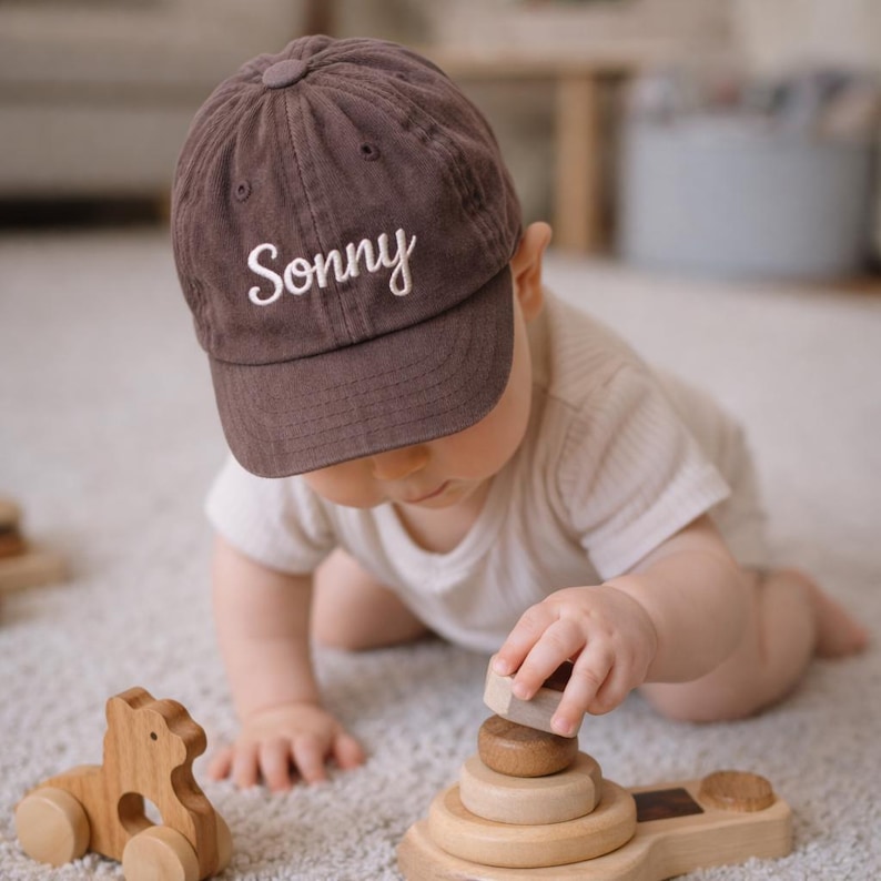 K&ouml;nnte beinhalten: Braune Baseballkappe mit dem Namen "Sonny" in Wei&szlig; gestickt. Ein Baby tr&auml;gt die Kappe und spielt mit Holzspielzeug, darunter ein Stapelring und ein Holztier. Die Szene spielt sich auf einem hellfarbigen Teppich ab.