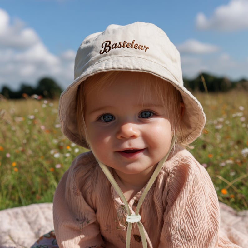 Pu&ograve; includere: Un bambino sorridente indossa un cappello da sole beige con la scritta "Basteleur" ricamata in marrone. Il cappello ha un sottogola. Il bambino indossa un vestito rosa ed &egrave; seduto in un campo di fiori selvatici.