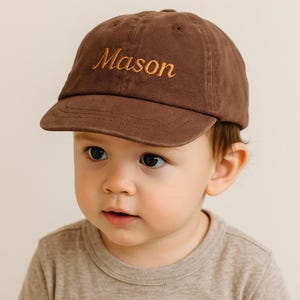 May include: A brown baseball cap embroidered with the name "Mason" in gold. The cap features a curved peak and a traditional six-panel construction. The cap is made from a soft, durable material.