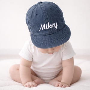 May include: A blue denim baseball cap with the name "Mikey" embroidered in white script. The cap is worn by a baby wearing a white onesie. The baby is sitting on a white surface.
