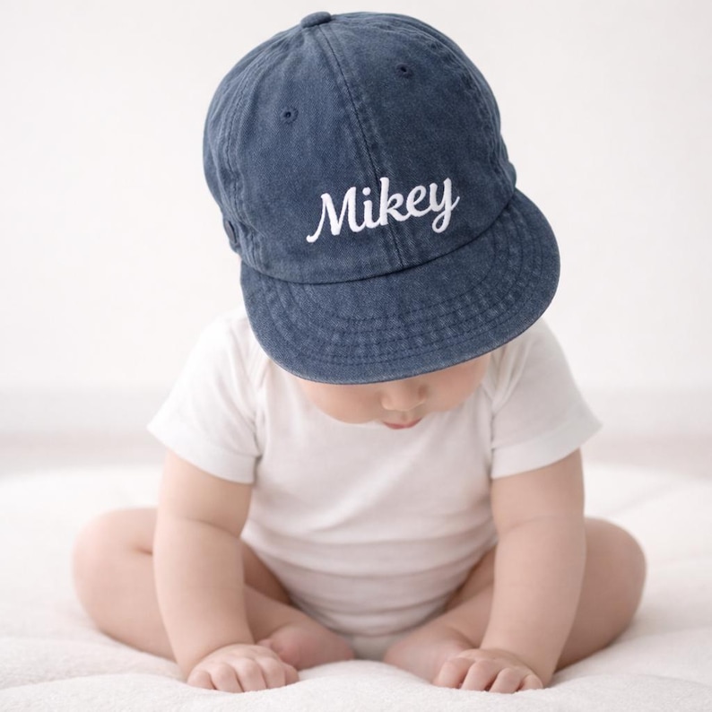 K&ouml;nnte beinhalten: Eine blaue Jeans-Baseballkappe mit dem Namen "Mikey" in wei&szlig;er Schrift gestickt. Die Kappe wird von einem Baby getragen, das einen wei&szlig;en Body tr&auml;gt. Das Baby sitzt auf einer wei&szlig;en Oberfl&auml;che.