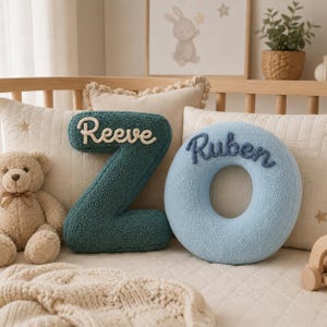 May include: A dark teal letter Z-shaped pillow with the name Reeve in white script and a light blue circular O-shaped pillow with the name Ruben in dark blue script. Both pillows are made of a textured, fuzzy fabric and are placed together.