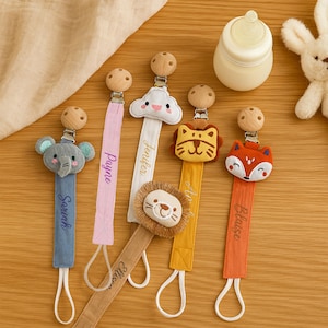 Op de afbeelding: Een verzameling fopspeenclips voor baby's met houten clips en stoffen bandjes. Elke clip heeft een ander dierontwerp: een olifant, een wolk, een leeuw en een vos. De namen "Sarah", "Payne", "Jenifer", "Allison" en "Blaise" zijn op de bandjes geborduurd.