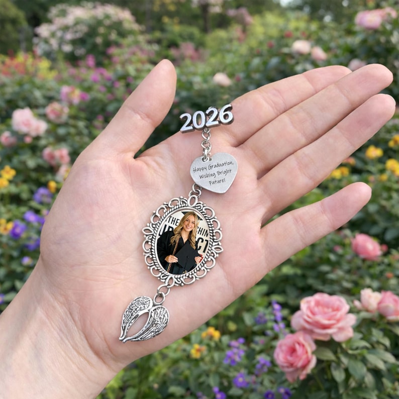 Pu&ograve; includere: Un ornamento di laurea argentato con una foto di una laureata, l'anno 2026 e il testo "Happy Graduation, Wishing Bright Future!". L'ornamento ha un design a cuore e ali.