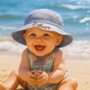 May include: A smiling baby wearing a blue and white striped sun hat with the name "Ethan" embroidered on the front. The baby is wearing a floral print swimsuit and sitting on a sandy beach with the ocean in the background.