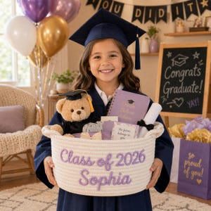 May include: A young graduate in a navy blue cap and gown holds a woven basket filled with graduation gifts. The basket is embroidered with "Class of 2026 Sophia." A teddy bear, notebook, and diploma are visible. Balloons and a chalkboard with "Congrats Graduate!" are in the background.