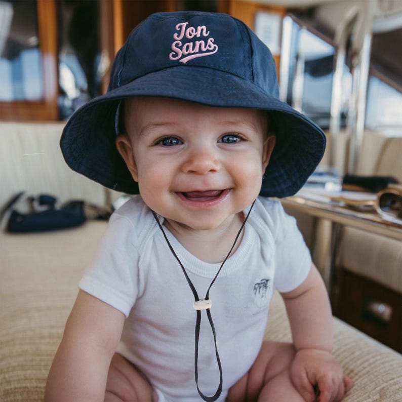 Pu&ograve; includere: Un bambino sorridente con un cappello da sole blu navy con la scritta "Jon Sans" in corsivo rosa. Il bambino indossa anche un body bianco con un cordino nero intorno al collo.