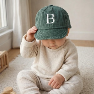 May include: A green baseball cap with the letter "B" embroidered in white. The cap is worn by a baby wearing a cream-colored sweater and pants. The baby is sitting on a white rug.