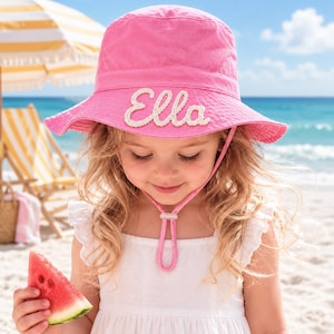 Puede incluir: Un sombrero de pescador de color rosa brillante con el nombre Ella bordado en letras blancas cursivas en la parte delantera. El sombrero incluye una correa de barbilla rosa a juego con un cierre ajustable para asegurar el sombrero al usarlo.