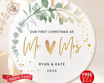 Könnte beinhalten: Ein rundes, weißes Keramikornament mit einem goldenen Band zum Aufhängen. Das Design zeigt einen Kranz aus grünen und goldenen Eukalyptusblättern, den Text "OUR FIRST CHRISTMAS AS Mr ♥ Mrs", die Namen "RYAN & KATE" und das Jahr "2025" in Schwarz und Gold.