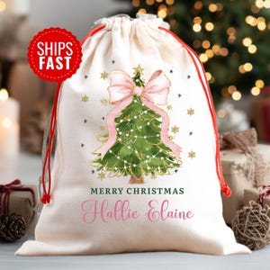 K&ouml;nnte beinhalten: Ein cremefarbener Stoffbeutel mit Kordelzug, verziert mit einer gr&uuml;nen Aquarell-Illustration eines Weihnachtsbaums, der von einer gro&szlig;en rosa Schleife gekr&ouml;nt wird. Der Text Merry Christmas und der Name Hallie Elaine sind darunter gedruckt.