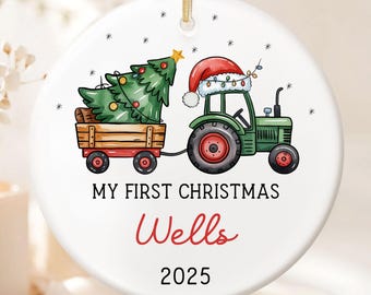 Könnte beinhalten: Runder Weihnachtsschmuck mit einem grünen Traktor, der einen Holzkarren mit einem Weihnachtsbaum zieht. Der Traktor hat einen Weihnachtsmannhut und Lichter. Der Text lautet "MY FIRST CHRISTMAS Wells 2025".