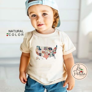 May include: A toddler wearing a natural colored t-shirt with a patchwork American flag design. The shirt has short sleeves and a crew neck. The toddler is also wearing a light blue baseball cap and blue jeans.