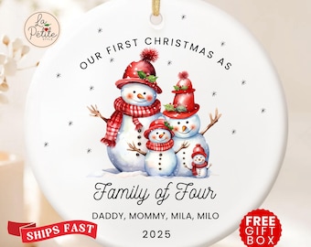 May include: A white ceramic Christmas ornament featuring a family of snowmen wearing red hats and scarves. The text "Our First Christmas As Family of Four" is displayed, along with the names and the year 2025.