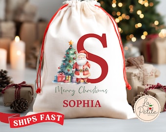 May include: Cream-colored Christmas gift bag with a red drawstring. The bag features a watercolor illustration of Santa Claus, a Christmas tree, and presents. A large red letter "S" and the name "Sophia" are printed on the bag, along with the words "Merry Christmas".