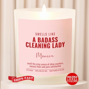May include: A white candle in a glass jar with a light pink label featuring red text that reads: SMELLS LIKE A BADASS CLEANING LADY, Monica, smell the crisp aroma of shiny counters, vacuum lines and pure satisfaction, 8 OZ VEGAN, 100% PURE SOY WAX.