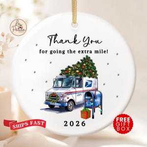 May include: A round white ceramic ornament with a gold hanging ribbon. It features a graphic of a USPS mail truck carrying a Christmas tree, a blue mailbox, and a gift. Text reads: Thank You for going the extra mile! and the year 2026.
