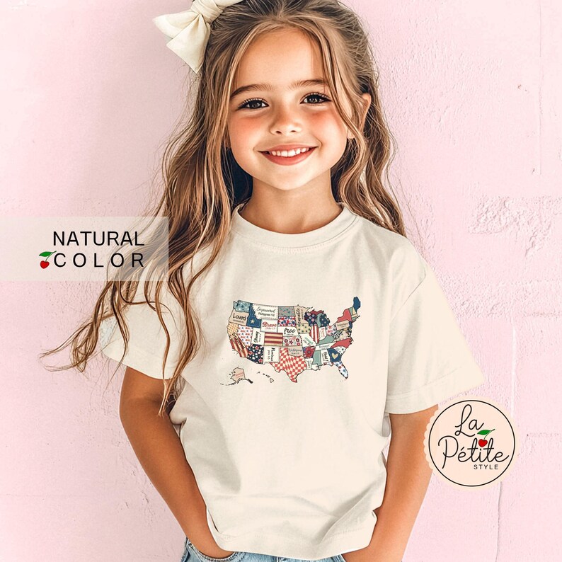 May include: A young girl wearing a white t-shirt with a patchwork design of the United States of America. The shirt has a natural color and the design is in red, white, and blue. The girl has long brown hair and is wearing a white bow in her hair.