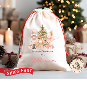 May include: A cream-colored Christmas gift bag with a red drawstring closure. The bag features a watercolor illustration of a nutcracker, a Christmas tree, a ballerina, and gifts. The text "Special Delivery For Addison" is printed on the bag.