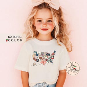 May include: A young girl wearing a white t-shirt with a patchwork American flag design. The shirt has the text "La Petite Style" on the bottom right corner.
