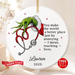 May include: A white ceramic ornament with a Grinch-themed design. It features a green hand, a red stethoscope in a heart shape, and the text "You make the world a better place just by annoying - I mean, touching - lives." The name "Lauren" and the year "2025" are also included.