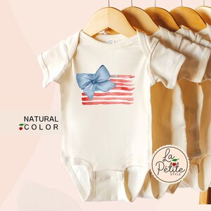 May include: A white baby onesie with a watercolor American flag design and a blue bow. The onesie has the text "La Petite Style" on the bottom right corner.
