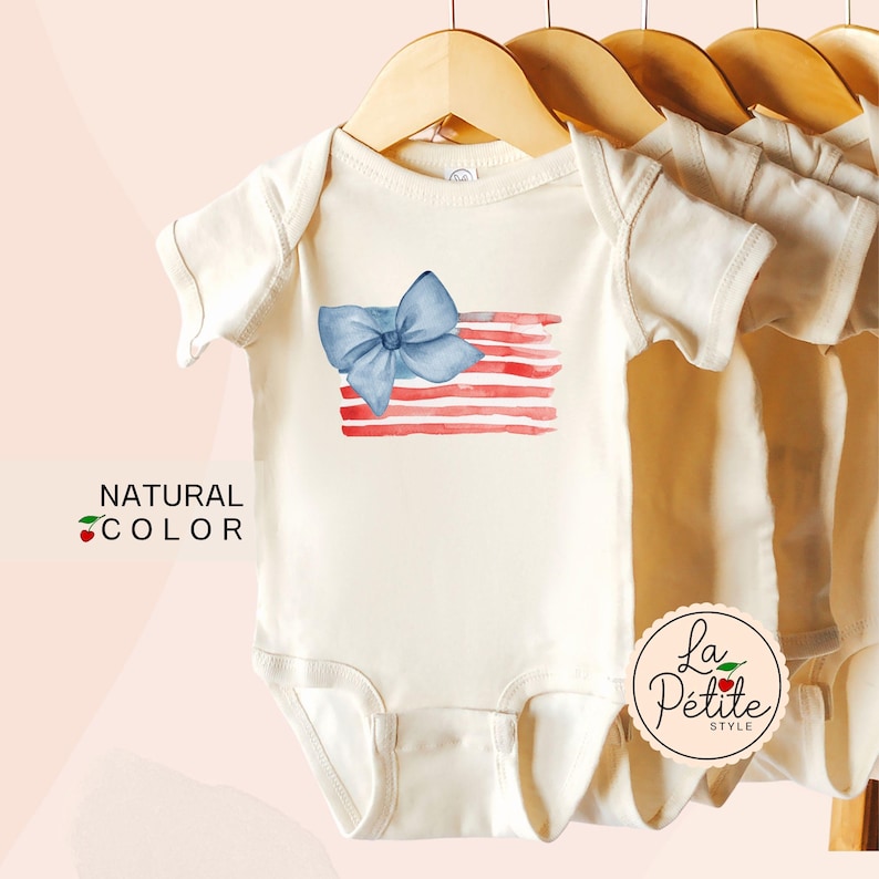 May include: A white baby onesie with a watercolor American flag design and a blue bow. The onesie has the text "La Petite Style" on the bottom right corner.
