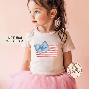 May include: A young girl wearing a pink t-shirt with a blue bow and red and white watercolor stripes. The shirt has the text "NATURAL COLOR" on the left side. The girl is wearing a pink tulle skirt.