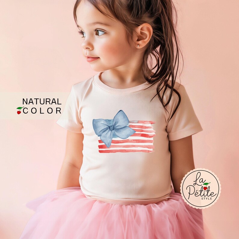 May include: A young girl wearing a pink t-shirt with a blue bow and red and white watercolor stripes. The shirt has the text "NATURAL COLOR" on the left side. The girl is wearing a pink tulle skirt.