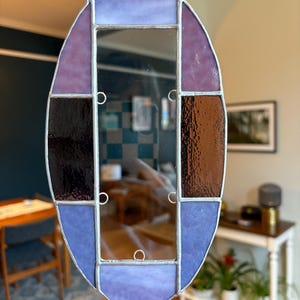 May include: An oval-shaped stained glass mirror with a silver chain for hanging. The glass is a mix of purple, blue, and brown hues, with a rectangular mirror in the center. The design is geometric and features a variety of textures.