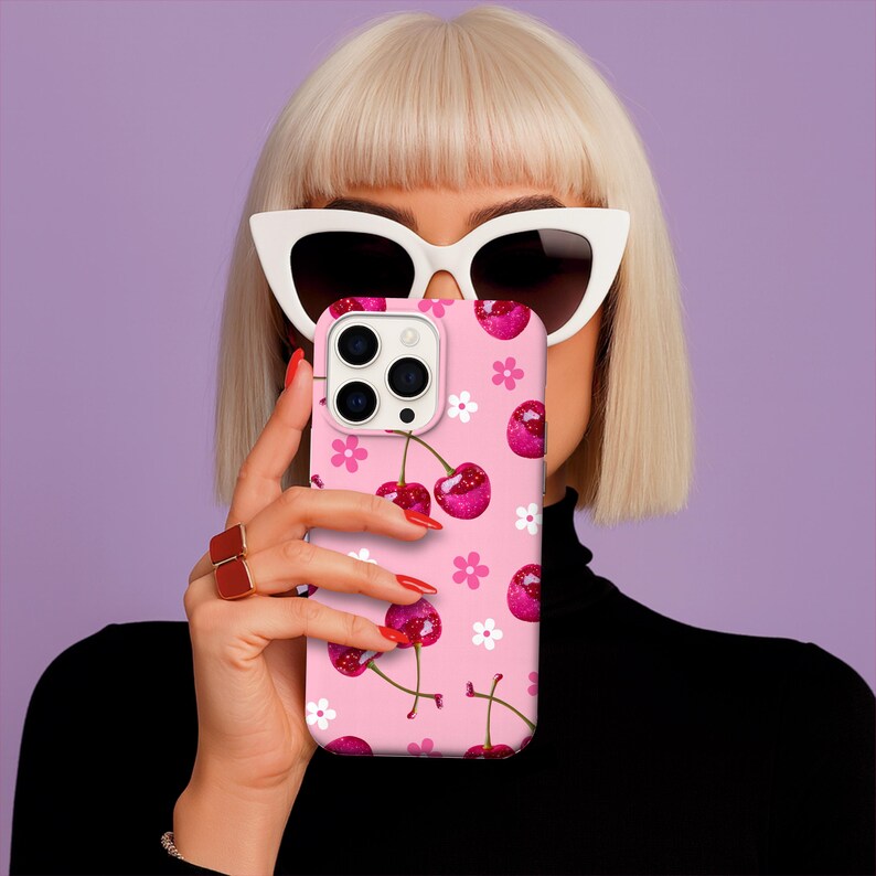 May include: Pink phone case with a cherry pattern. The case features a repeating design of red cherries and white flowers on a pink background. The phone case is held up in front of a person's face.