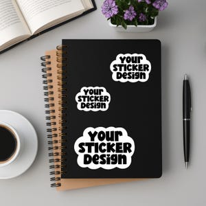 May include: A black spiral-bound notebook with three white stickers that read "Your Sticker Design" in a bold, rounded font. A pen, a cup of coffee, a book, and a small pot of purple flowers are also visible.
