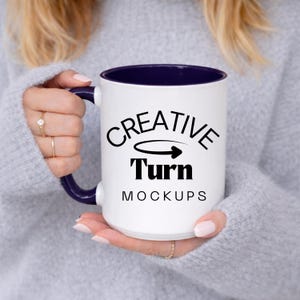 May include: White ceramic mug with a navy blue interior and handle. The mug features the text "CREATIVE Turn MOCKUPS" in black, with an arrow graphic. The mug is held by a person wearing a gray sweater.