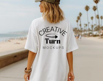 Op de afbeelding: Een wit t-shirt met de tekst "CREATIVE TURN MOCKUPS" in het zwart. De persoon op de afbeelding draagt een groene baseballcap en een bruine armband. De achtergrond is een strandscène met palmbomen.