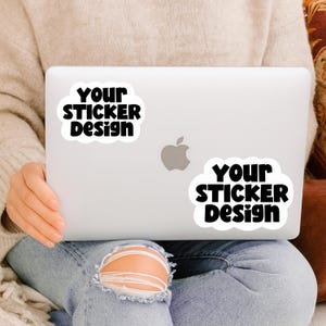 May include: A white laptop with two stickers that read "YOUR STICKER Design" in black text. The laptop has the Apple logo in the centre. The person is wearing a beige jumper and blue jeans.