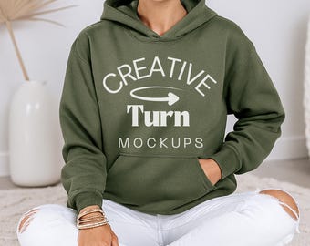 May include: An olive green hoodie with the text "CREATIVE Turn MOCKUPS" in white. The hoodie has a front pocket and a drawstring hood. The person in the image is wearing white ripped jeans and a gold bracelet.