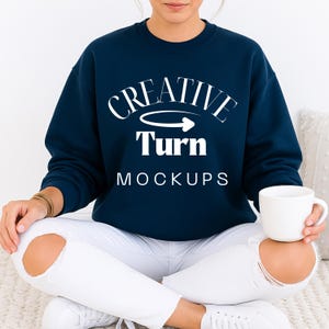 May include: A navy blue sweatshirt with the text "CREATIVE Turn MOCKUPS" in white. The sweatshirt is worn with white jeans. A white mug is held in the hand.