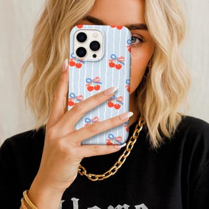 May include: A light blue phone case with a repeating pattern of red cherries tied with blue bows. The case has a vertical striped design and a camera cutout.