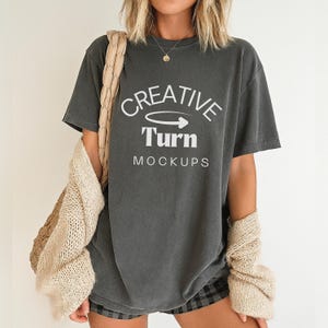 Może przedstawiać: Ciemnoszara koszulka z białym napisem "CREATIVE Turn MOCKUPS". Koszulka jest noszona z beżowym kardiganem i szortami w kratę. Koszulka jest makietą do projektowania graficznego.