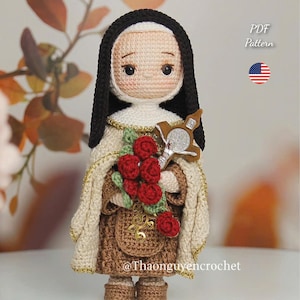 Puede incluir: Muñeca de ganchillo de Santa Teresa, con velo negro, toca blanca y hábito marrón. Sostiene una cruz y un ramo de rosas rojas. La muñeca tiene una capa con ribete dorado y una pequeña bolsa. El texto "St. Thérèse" está arriba.