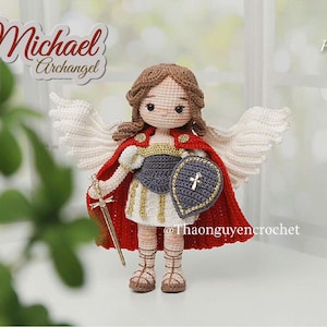 Puede incluir: Muñeca de ganchillo de Miguel Arcángel, con capa roja, armadura dorada y sosteniendo una espada y un escudo. La muñeca tiene alas blancas y cabello castaño. El texto "Michael Archangel" está arriba.